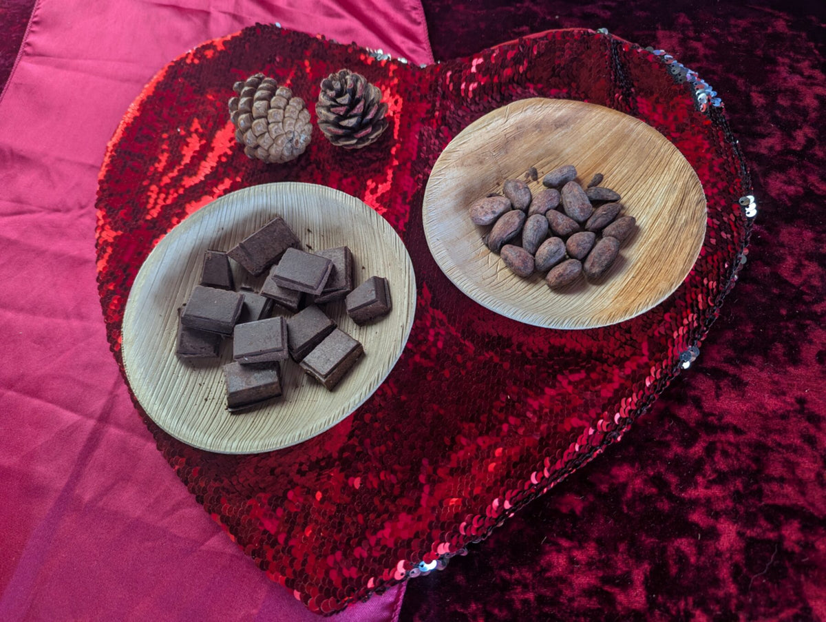 Squares of dark chocolate and roasted cacao beans on palm-leaf plates, on a red sequin heart